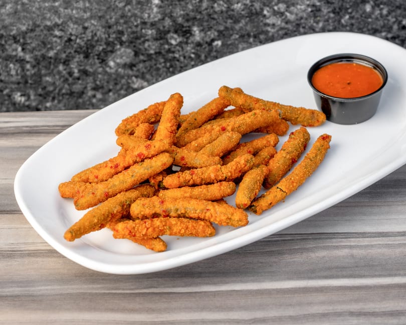A white platter filled with crispy fried pickle spears and a side of red dipping sauce.