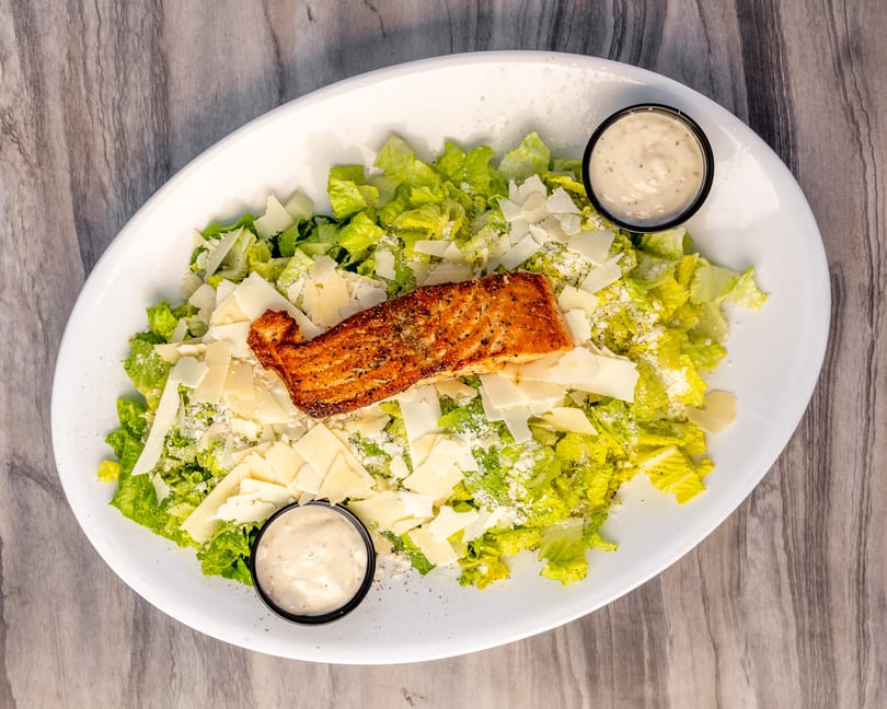 Grilled salmon Caesar salad with parmesan shavings and two cups of dressing on a white platter.