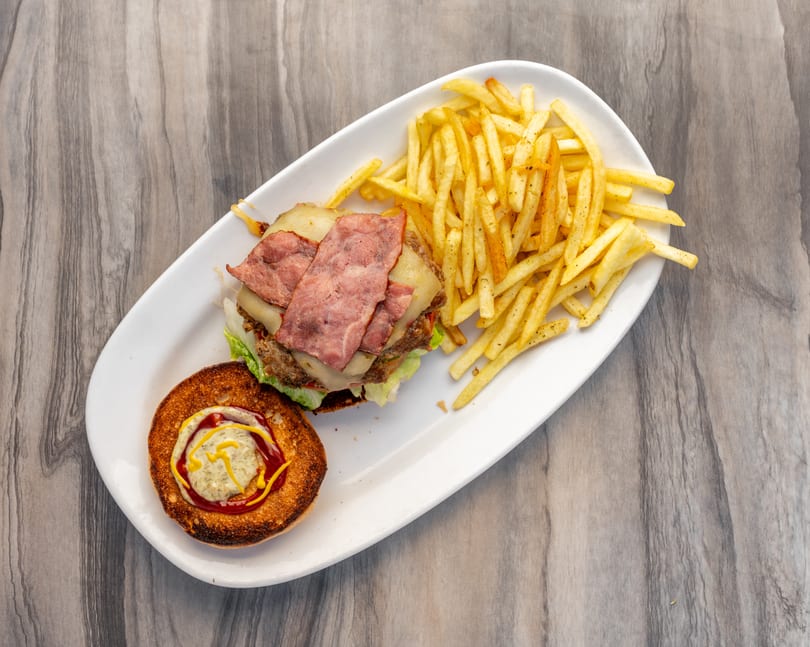 Overhead view of a bacon cheeseburger with fries on a white plate, bun top with condiments.