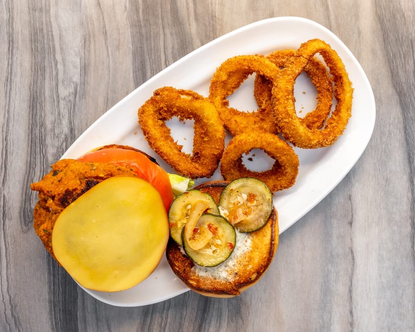 A deconstructed veggie burger with a patty, tomato, cheese, and zucchini on a bun, served with onion rings.
